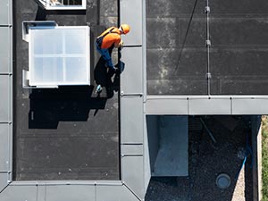 Construction worker in an orange hard hat and safety vest on a flat roof beside solar panels, secured by a safety line, seen from above.